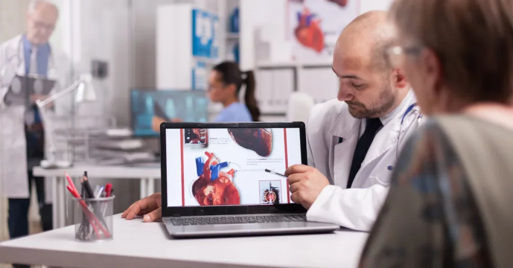 Cardiologista orientando paciente com imagens de um coração humano na tela de um notebook no consultório médico